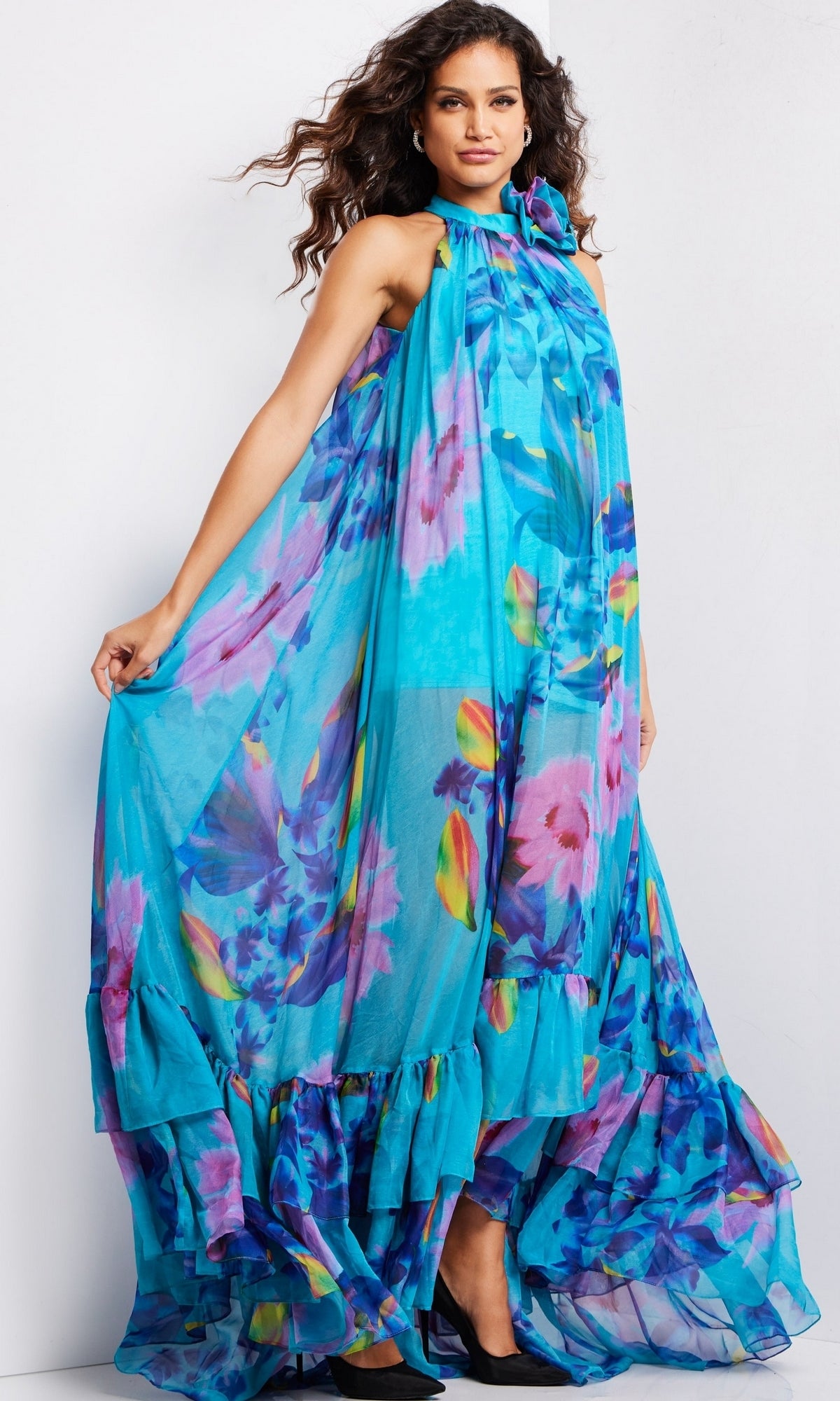 A woman stands against a plain background in the Long Prom Dress 38721 by Jovani—a sleeveless, floor-length turquoise gown featuring a colorful floral pattern and ruffled hem.